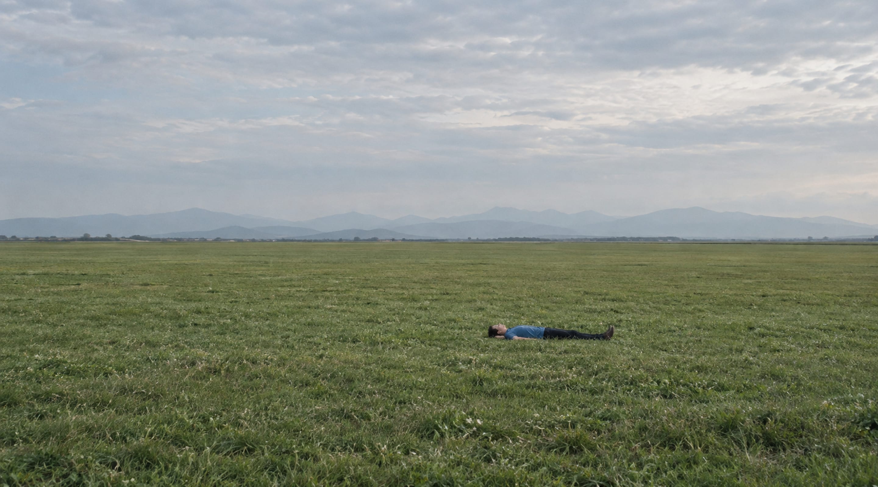 A person rests in an open field beneath distant hills and a pale sky.